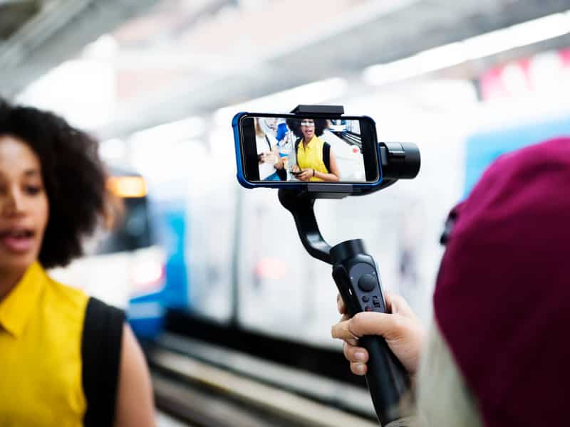 cameraman holding a smartphone on a selfie stick and shooting a woman with a yellow shirt